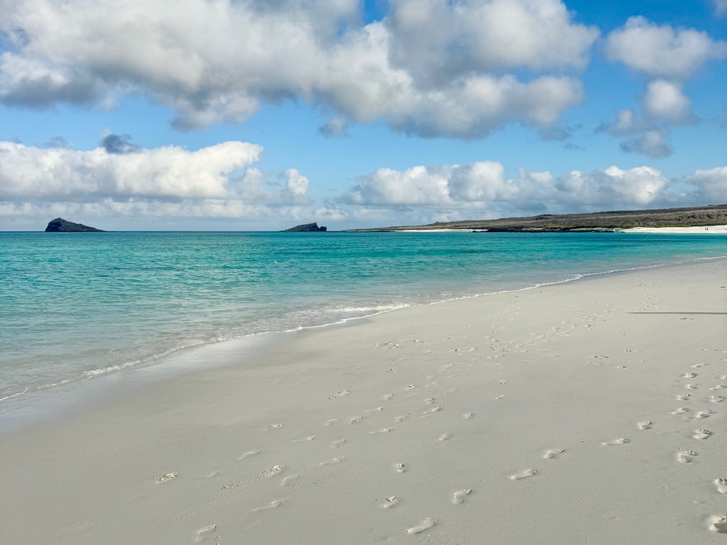 A serene beach scene with soft white sand and gentle turquoise waves under a partially cloudy blue sky, featuring distant rocky islets in the water.